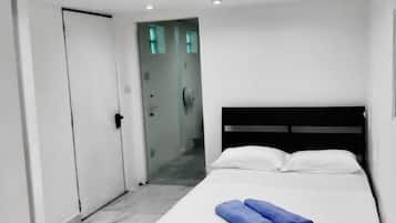 A modern hotel room with a large bed, white walls, and a glass door leading to a hallway.