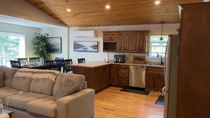 A cozy living room with a sofa and a dining area, a kitchen with wooden cabinets and a dishwasher, and a large window providing natural light. - Welcome to Sunset Cove, Cozy home, enjoy the landscaped property & outdoor area. (Goobies)