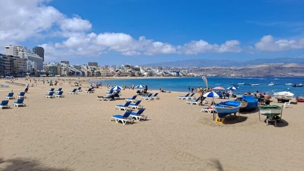 Ein Strand mit Liegestühlen und Sonnenschirmen, Booten sowie Gebäuden im Hintergrund.