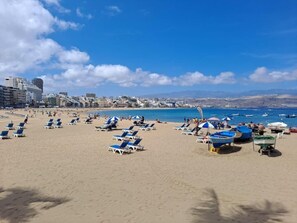 Una playa con tumbonas y sombrillas, barcos y edificios al fondo.