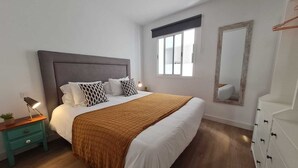 Ein Schlafzimmer mit einem großen Bett, einem Nachttisch mit Lampe, einem Schminktisch, einem Fenster mit Jalousien und einem Spiegel an der Wand.