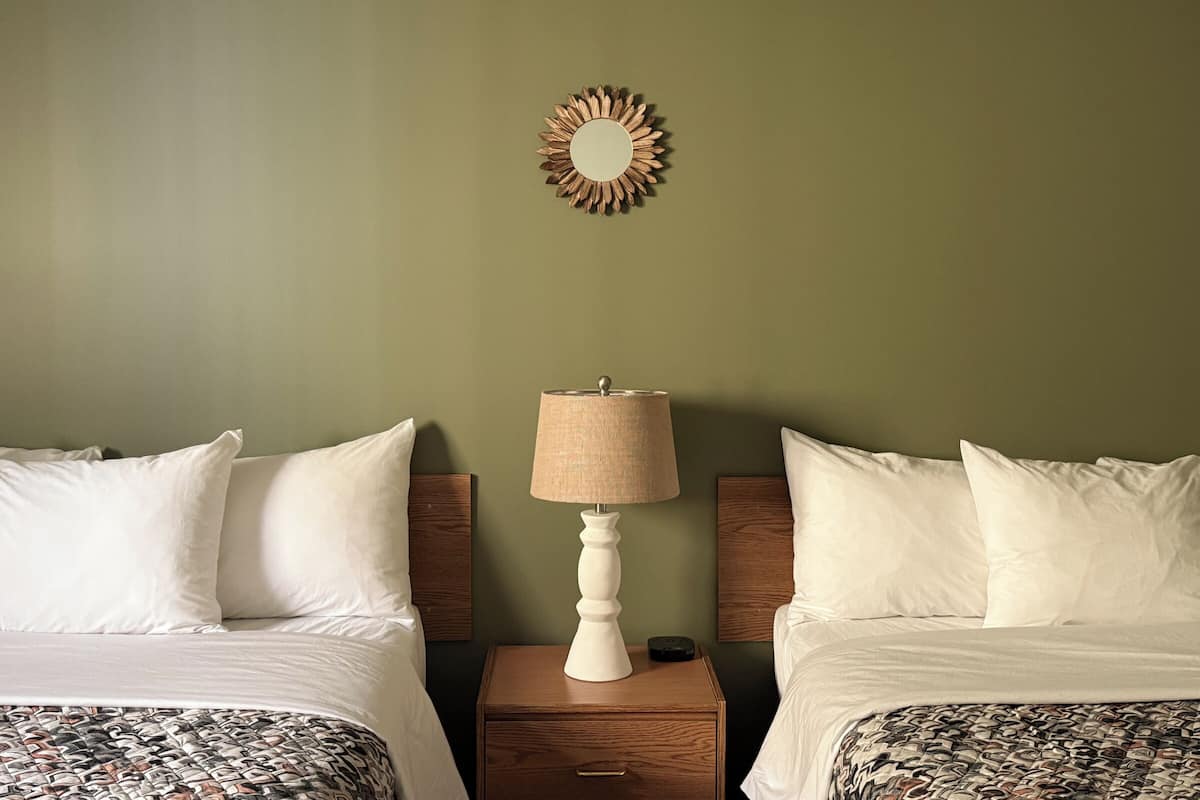 A hotel room with two beds, a nightstand with a lamp, and a wall-mounted mirror.