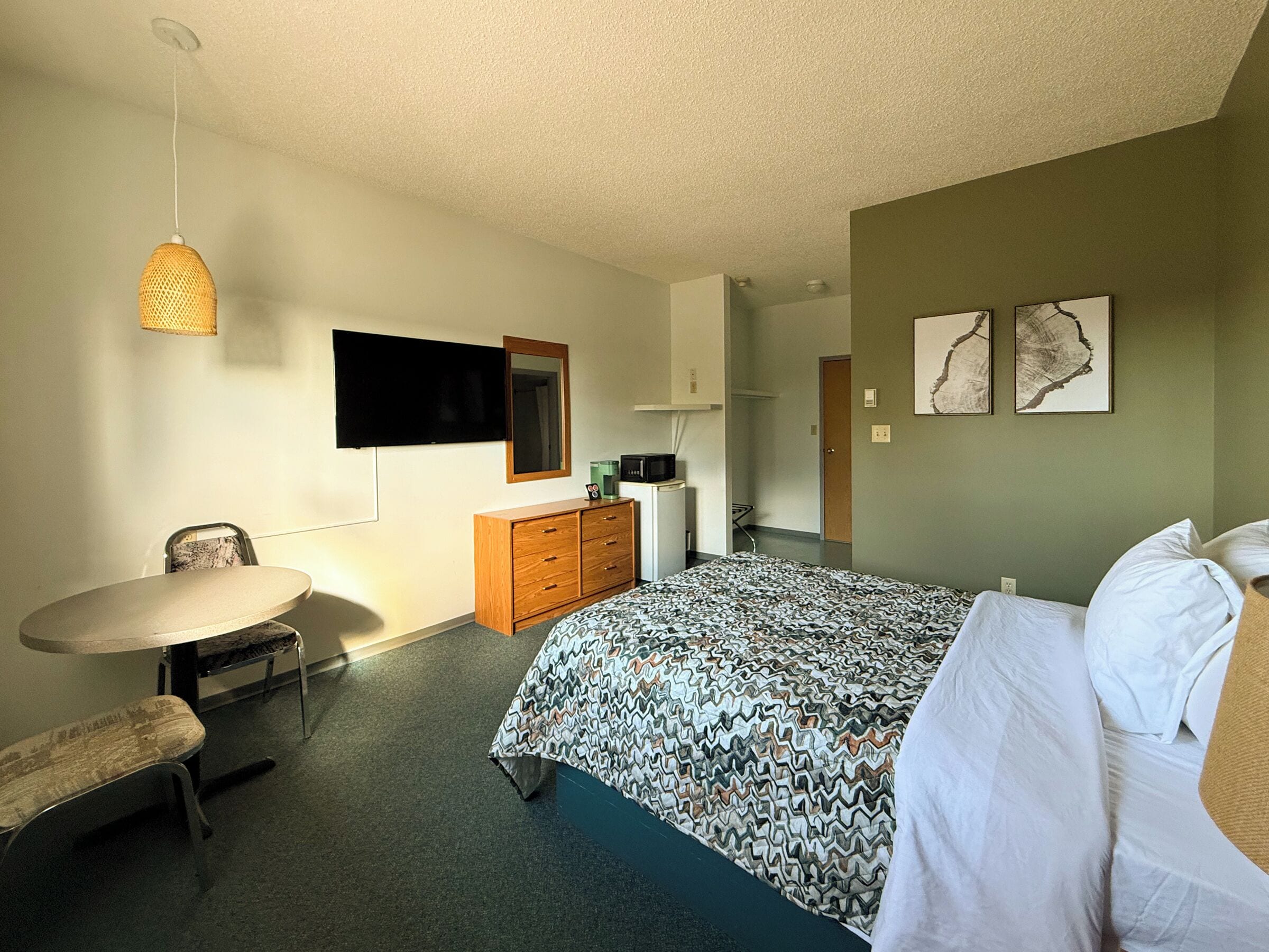 A hotel room with a bed, a desk, a chair, a dresser, a television, and artwork on the walls.