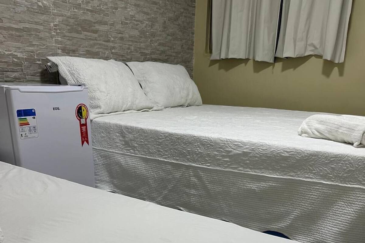 A hotel room with a bed, a white wall, a window with curtains, and a refrigerator with stickers.