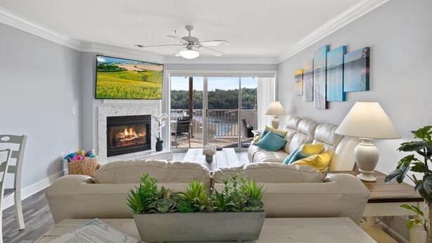 A living room with a fireplace, a ceiling fan, a sofa, a coffee table, and a view of the outdoors. - Lake View Condo - Heart of Osage Beach + Boat Slip (Osage Beach)
