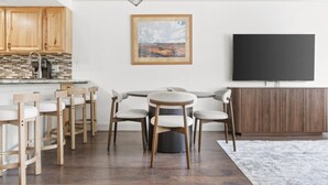 A modern kitchen with wooden cabinets, a tiled backsplash, and a built-in stove. There is a dining area with a round table and chairs, a flat-screen TV mounted on the wall, and a large window with a view of the outdoors. - The Alpine Estate At Copper Chase (Brian Head)