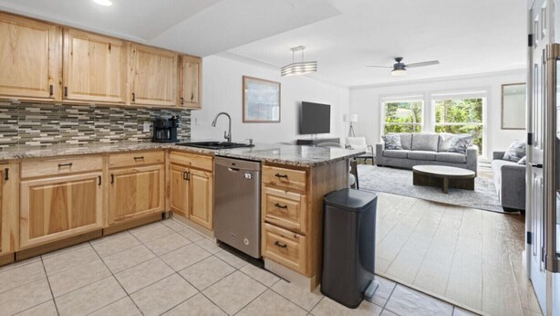 A modern kitchen with wooden cabinets, a dishwasher, and a large island. There is a dining area with a gray sofa and a coffee table. - The Alpine Estate At Copper Chase (Brian Head)