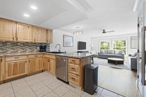 A modern kitchen with wooden cabinets, a dishwasher, and a large island. There is a dining area with a gray sofa and a coffee table. - The Alpine Estate At Copper Chase (Brian Head)