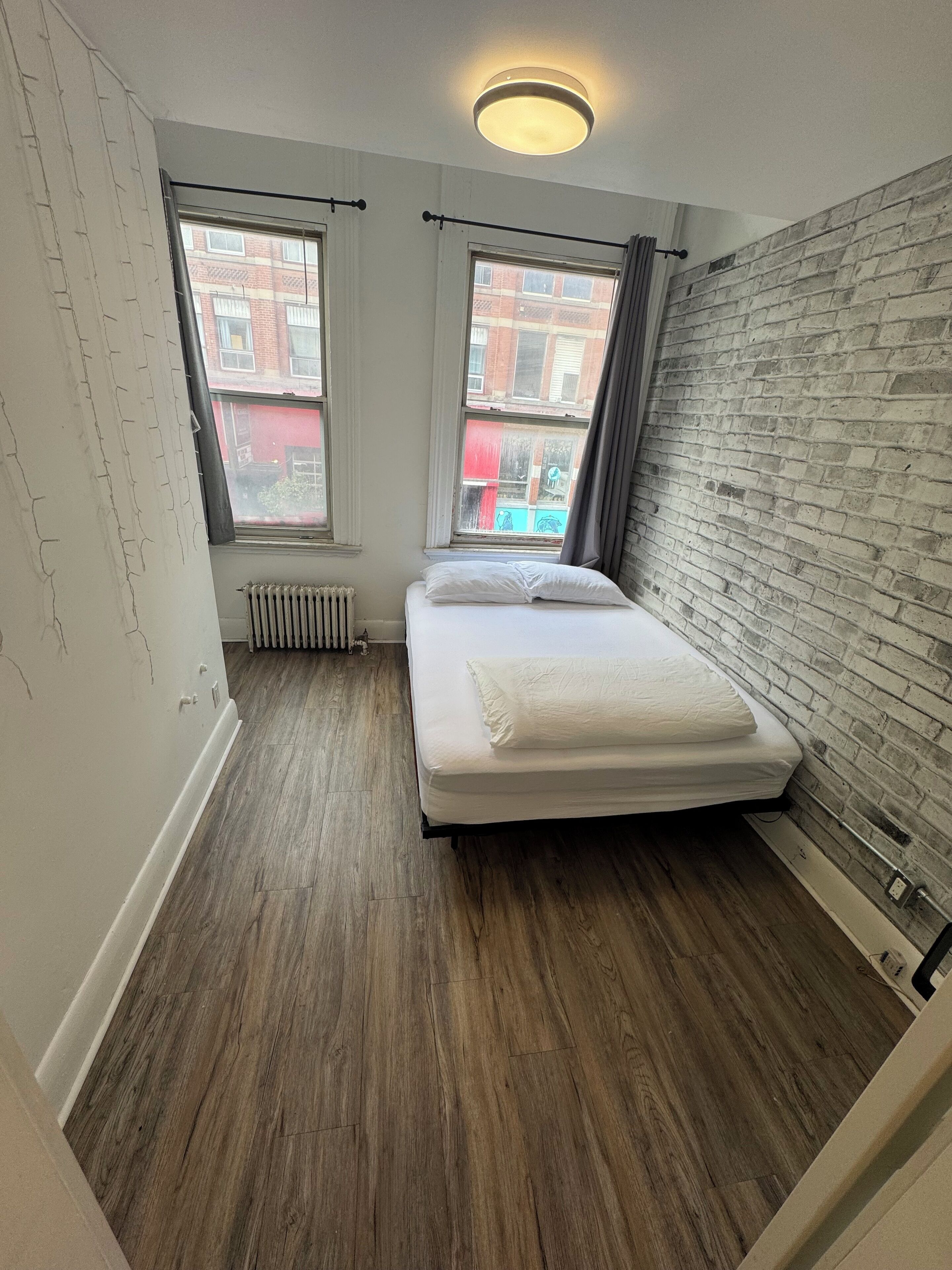 A small, modern bedroom with a brick wall, a wooden floor, a bed with white bedding, and two windows with curtains.