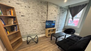 A modern living room with a brick wall, a black sofa, a wooden bookshelf, and a TV on a wooden stand. - Beautiful apartment on Richmond near via rail (London)