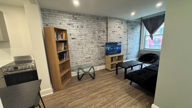 A living room with a brick wall, a wooden bookshelf, a TV, a black sofa, and a dining area with a table and chairs. - Beautiful apartment on Richmond near via rail (London)