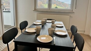 A dining table set with plates, cups, and cutlery in a room with a view of a hilly landscape through large windows. - Fitters' and vacation apartment near Heidelberg in the Odenwald with balcony & garden (Wilhelmsfeld)