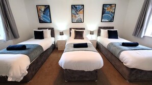 A hotel room with three single beds, each with a bedside table and lamp, and artwork on the walls.