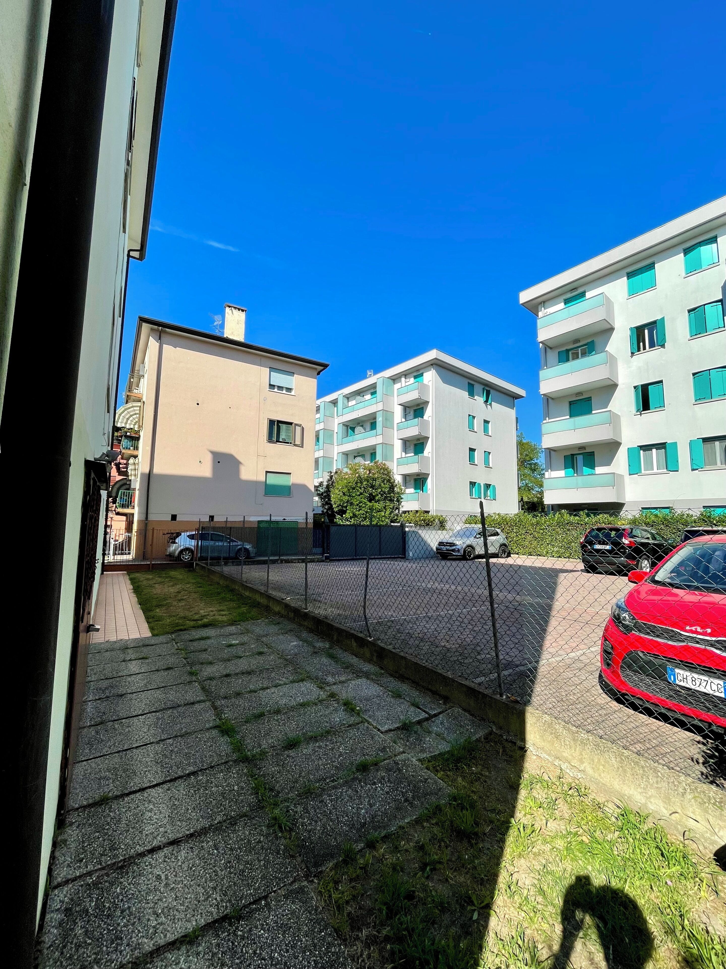 Ein Wohngebiet mit modernen Mehrfamilienhäusern, einem eingezäunten Parkplatz und einem roten Auto auf der Straße.