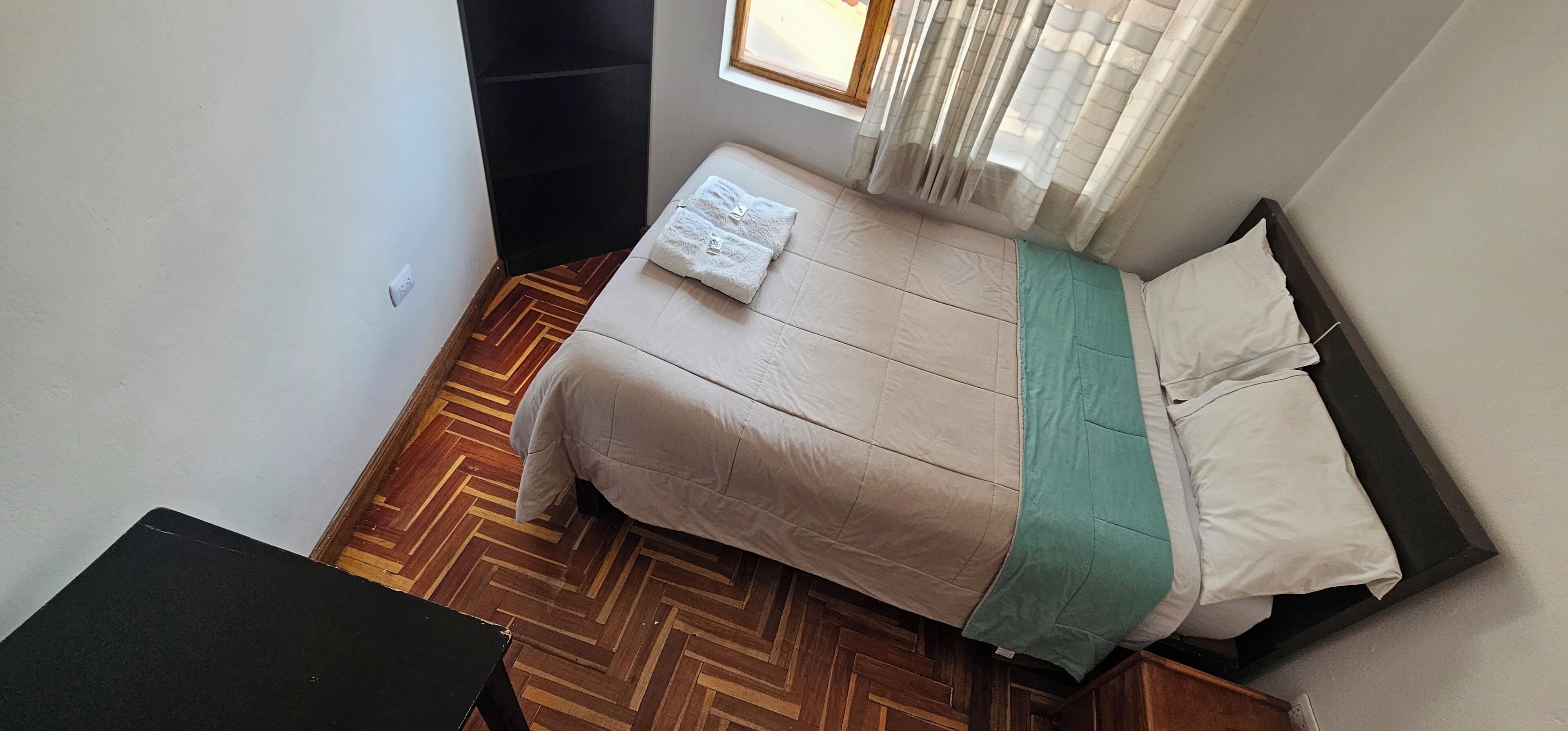 Un dormitorio pequeño y ordenado, con cama, una mesita y ventana con cortinas.