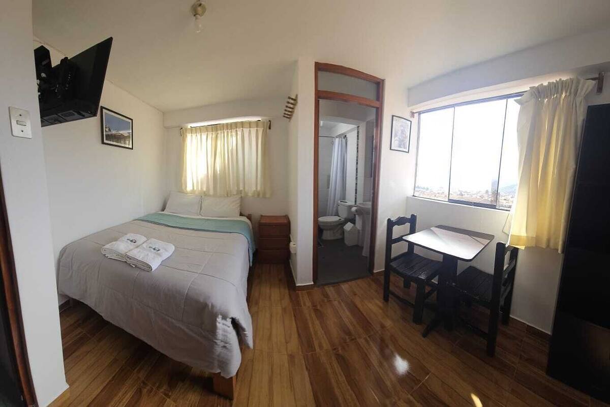 A hotel room with a bed, a desk, a chair, a TV, and a bathroom visible through an open door.