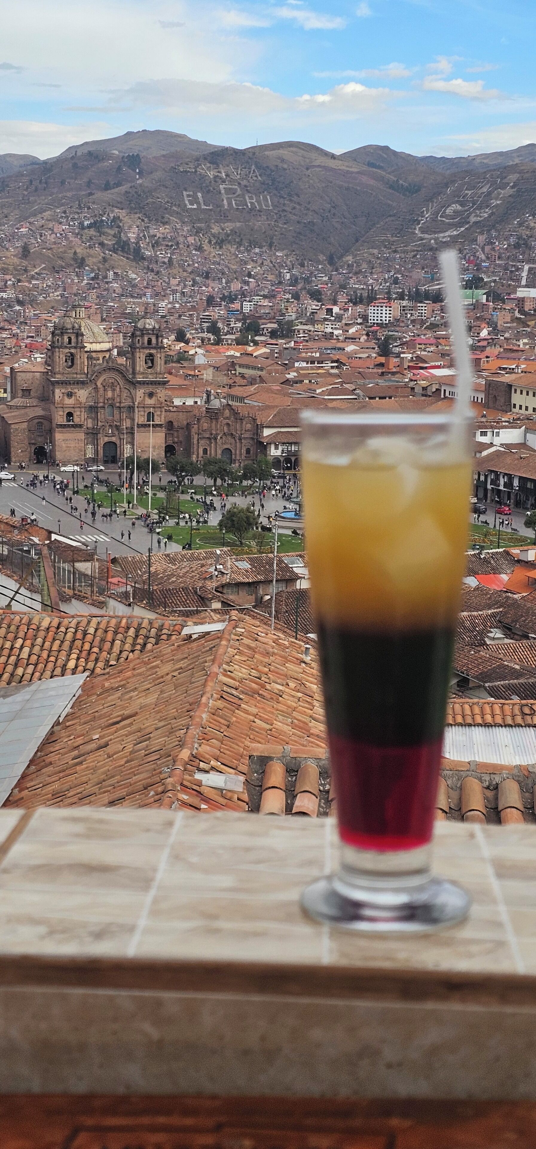 Um copo de bebida em uma varanda com vista para um horizonte urbano e arquitetura histórica, incluindo uma igreja imponente.