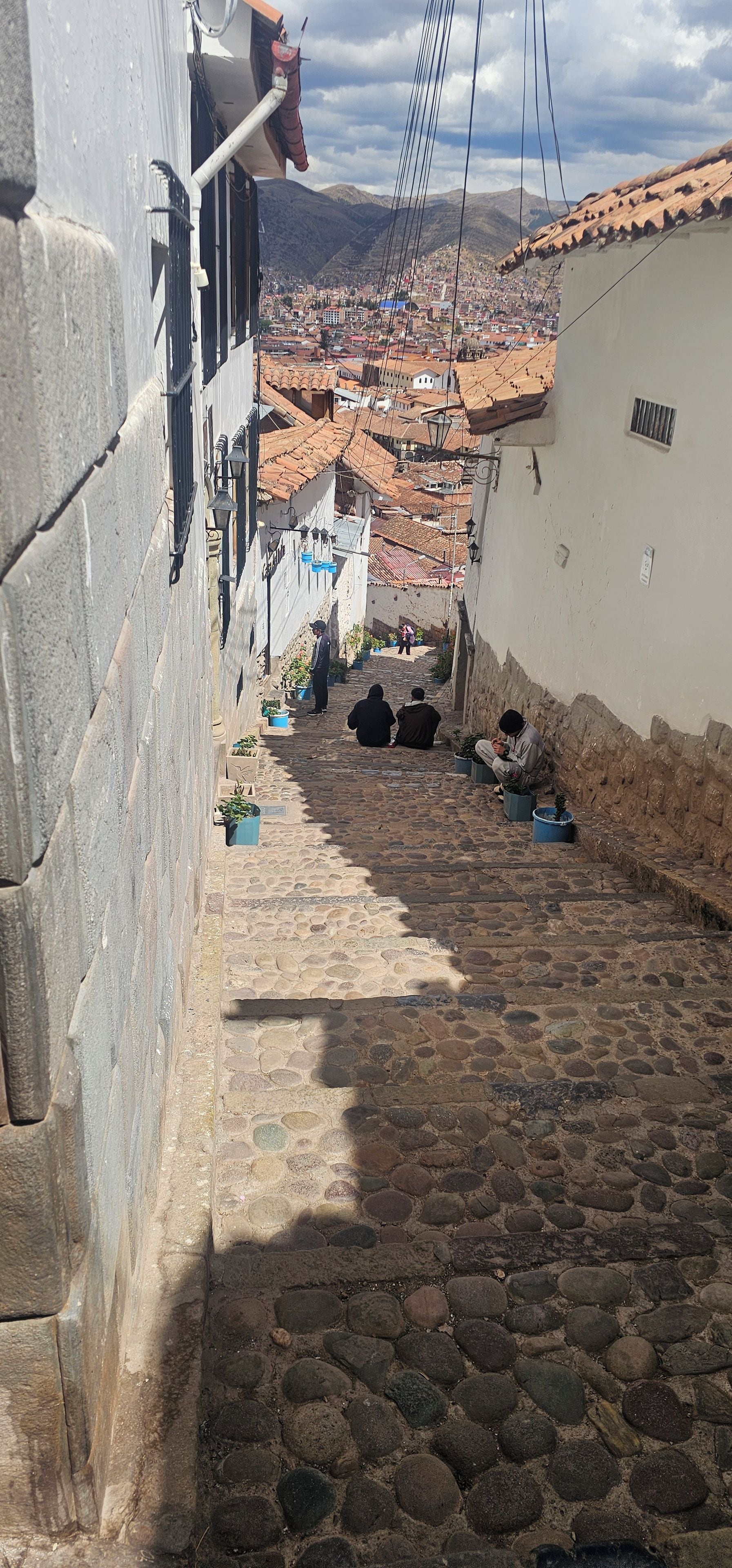 A cobblestone street with buildings on either side, a person sitting on the ground, and a view of a distant town.
