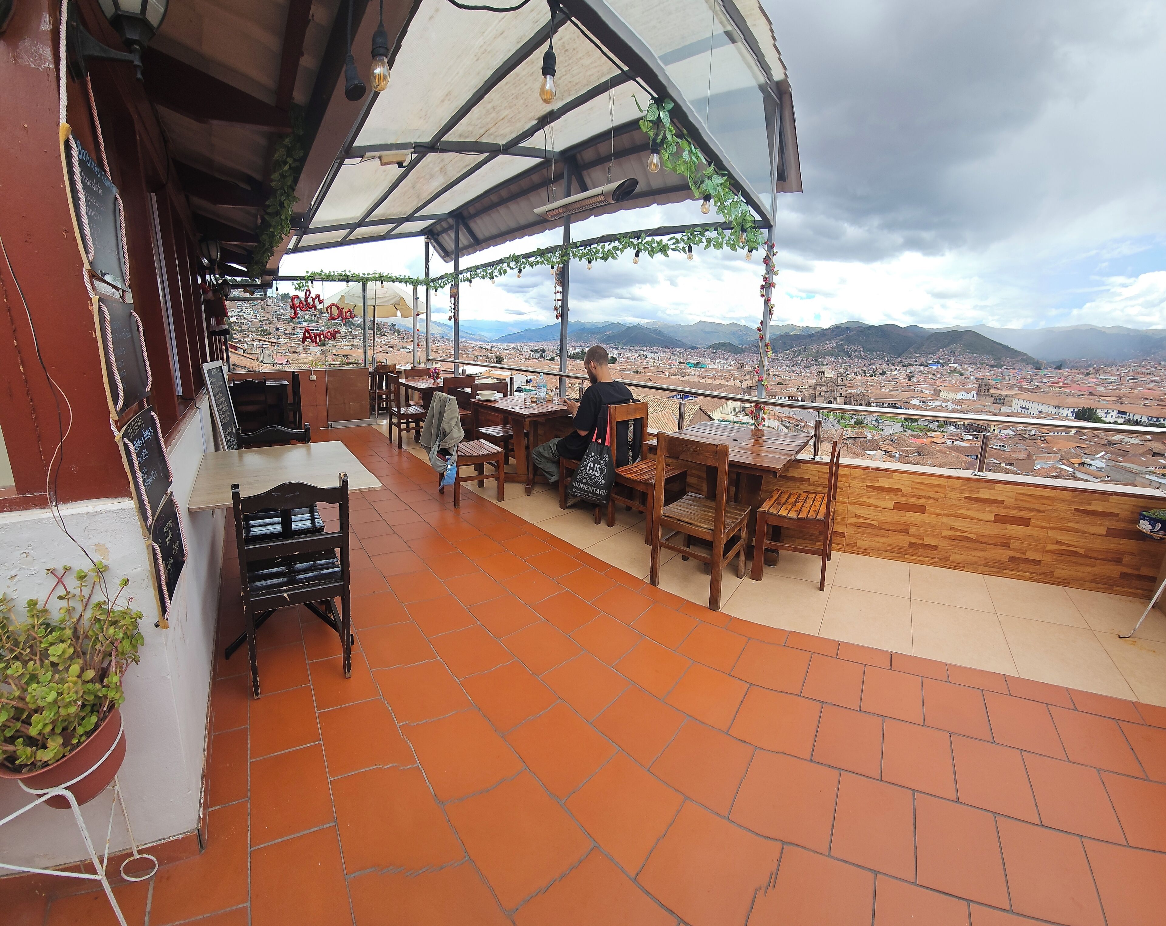 A terrace with wooden tables and chairs, a view of a cityscape, and a signboard.