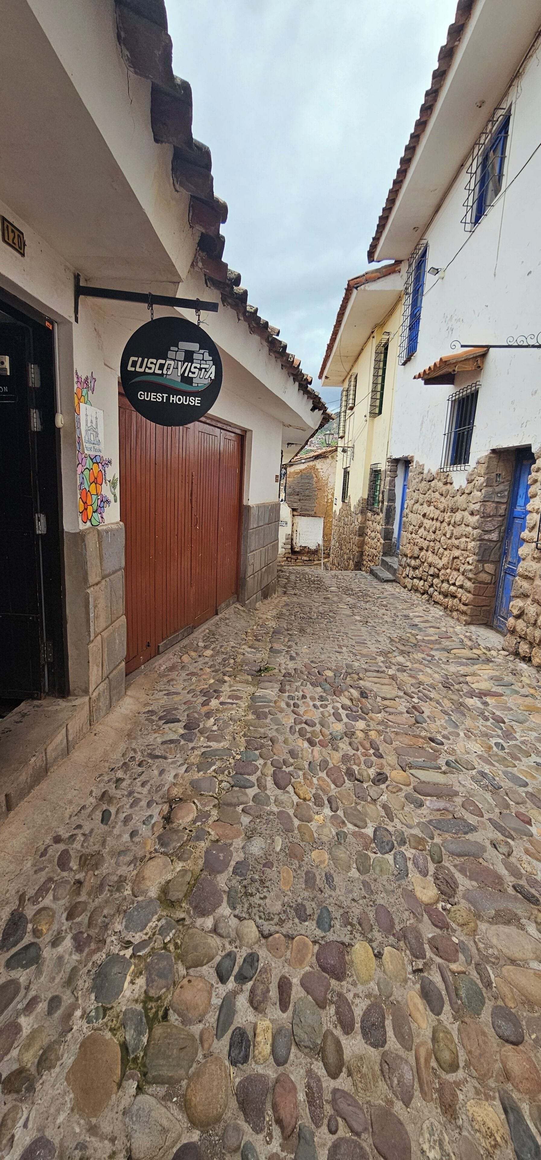 Une rue pavée bordée d’une maison d’hôtes appelée « CUSCO VISTA GUEST HOUSE » et d’un mur de pierre d’un côté.