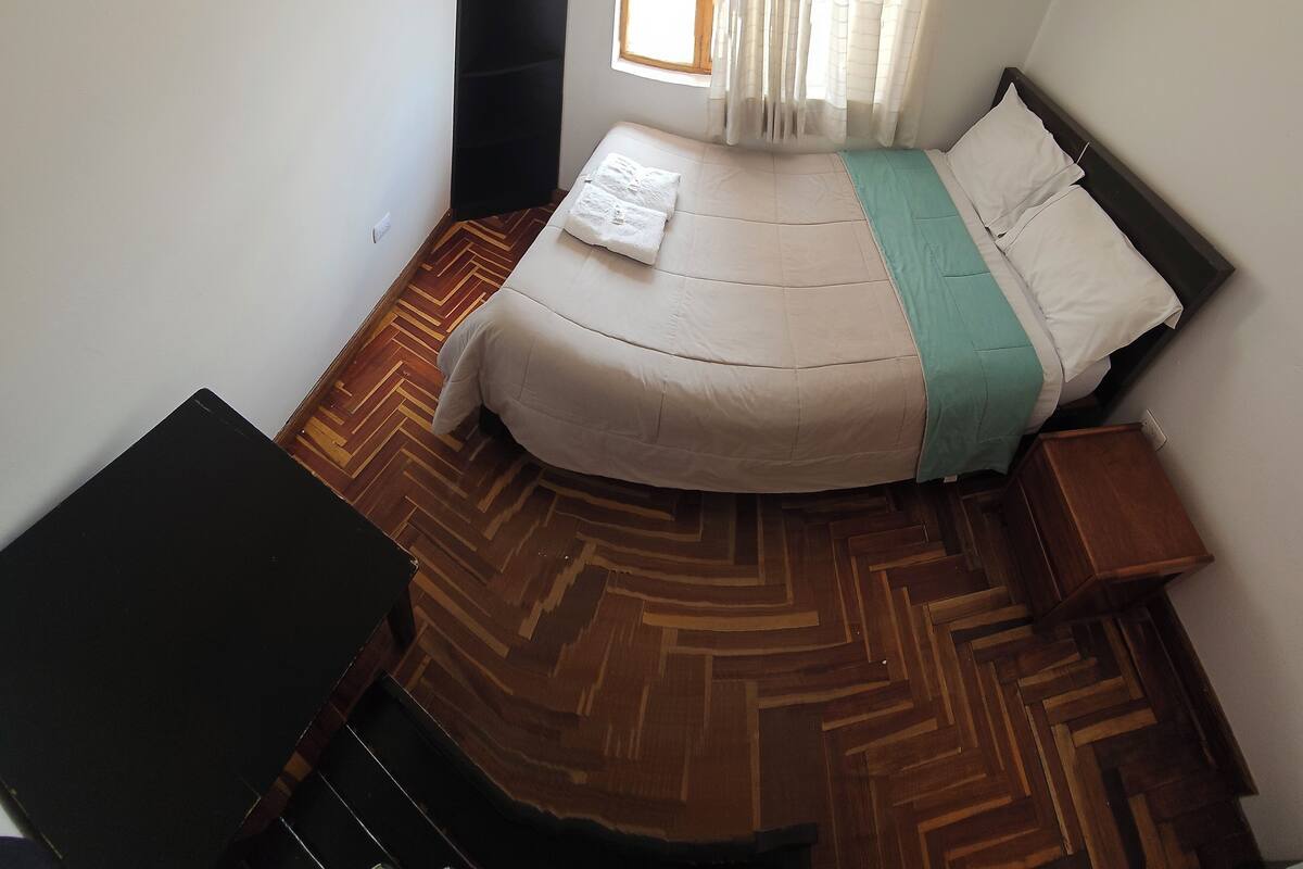 A bedroom with a bed, a wooden herringbone floor, a window with curtains, and a small bedside table.