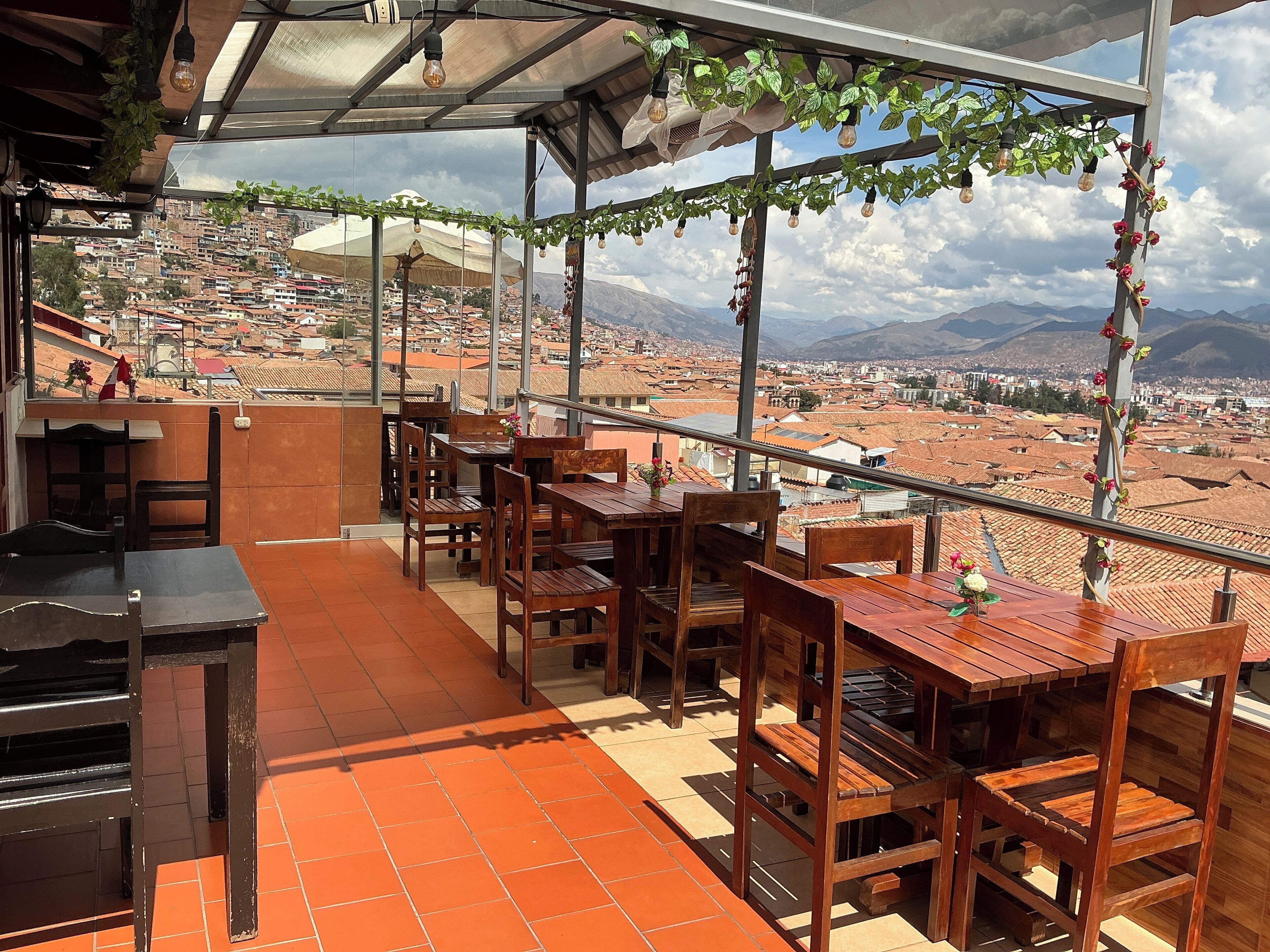 A terrace with wooden tables and chairs, a bar area, and a view of a cityscape with mountains in the background.