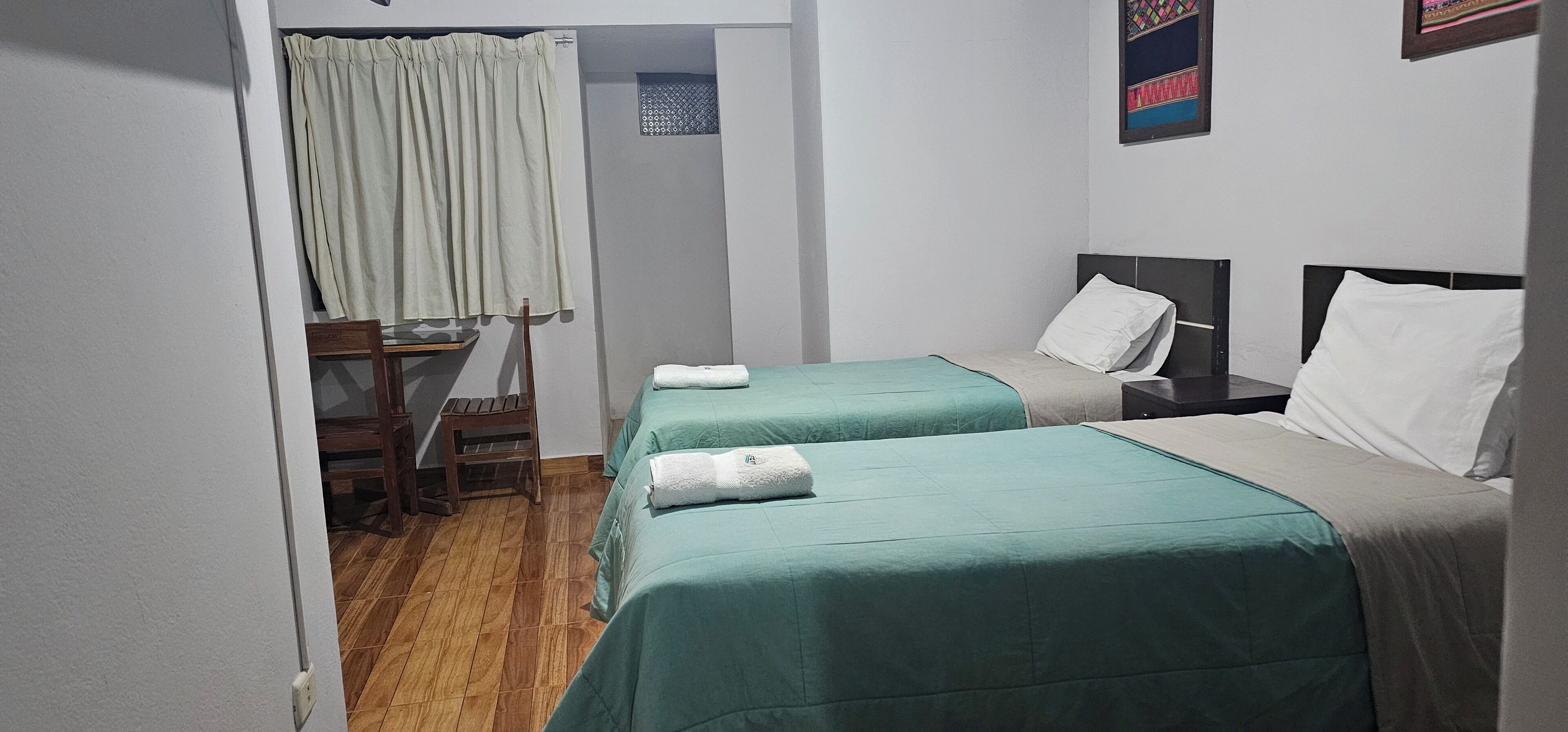 Habitación de hotel con dos camas individuales, una silla de madera, una mesita y una ventana con cortinas.