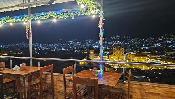 A rooftop restaurant with wooden tables and chairs, decorated with string lights and flowers. A cityscape with illuminated buildings is visible in the background. - CUSCO VISTA GUETS HOUSE (Cusco)