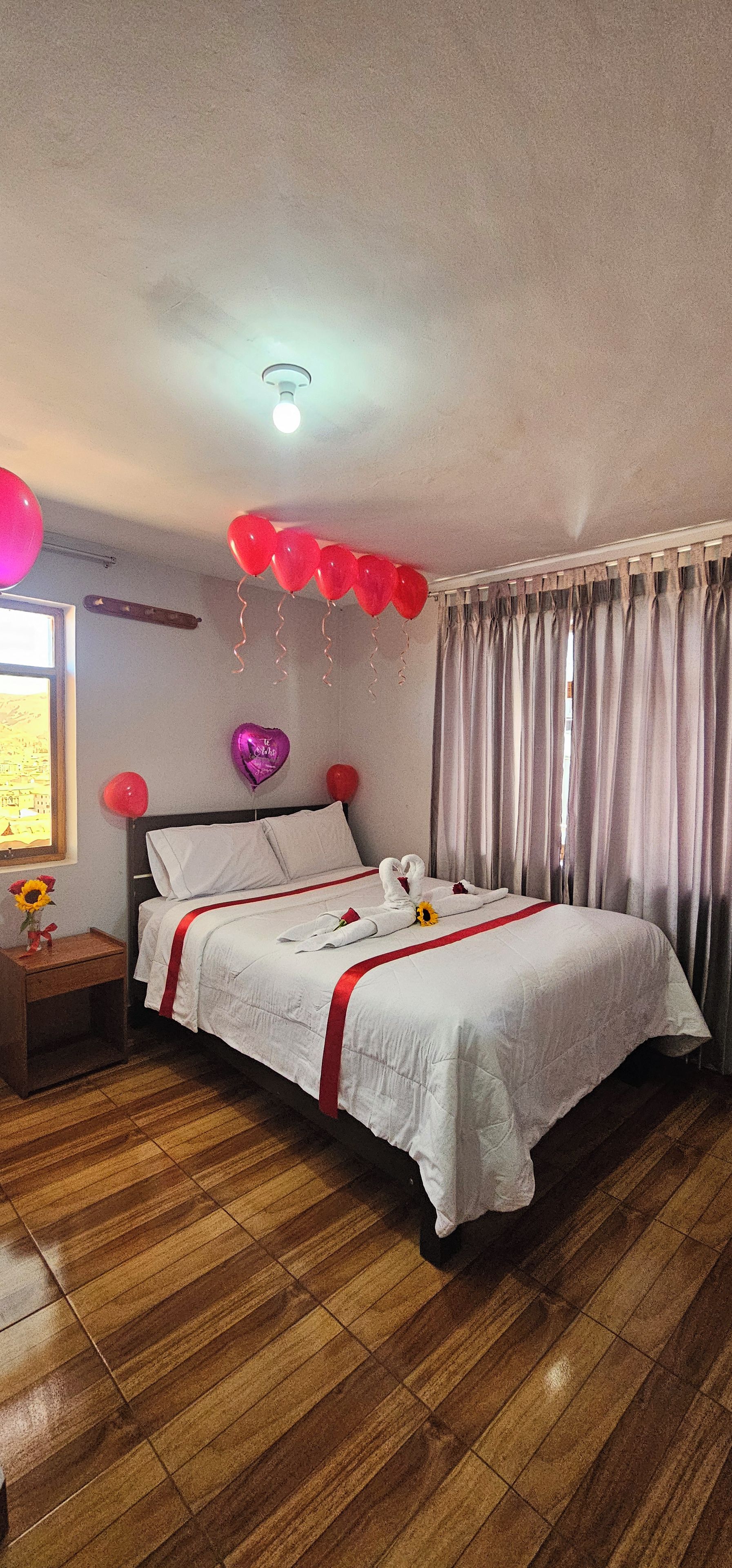 A hotel room with a wooden floor, a bed with white bedding and red accents, a nightstand with a vase of flowers, and a window with curtains.