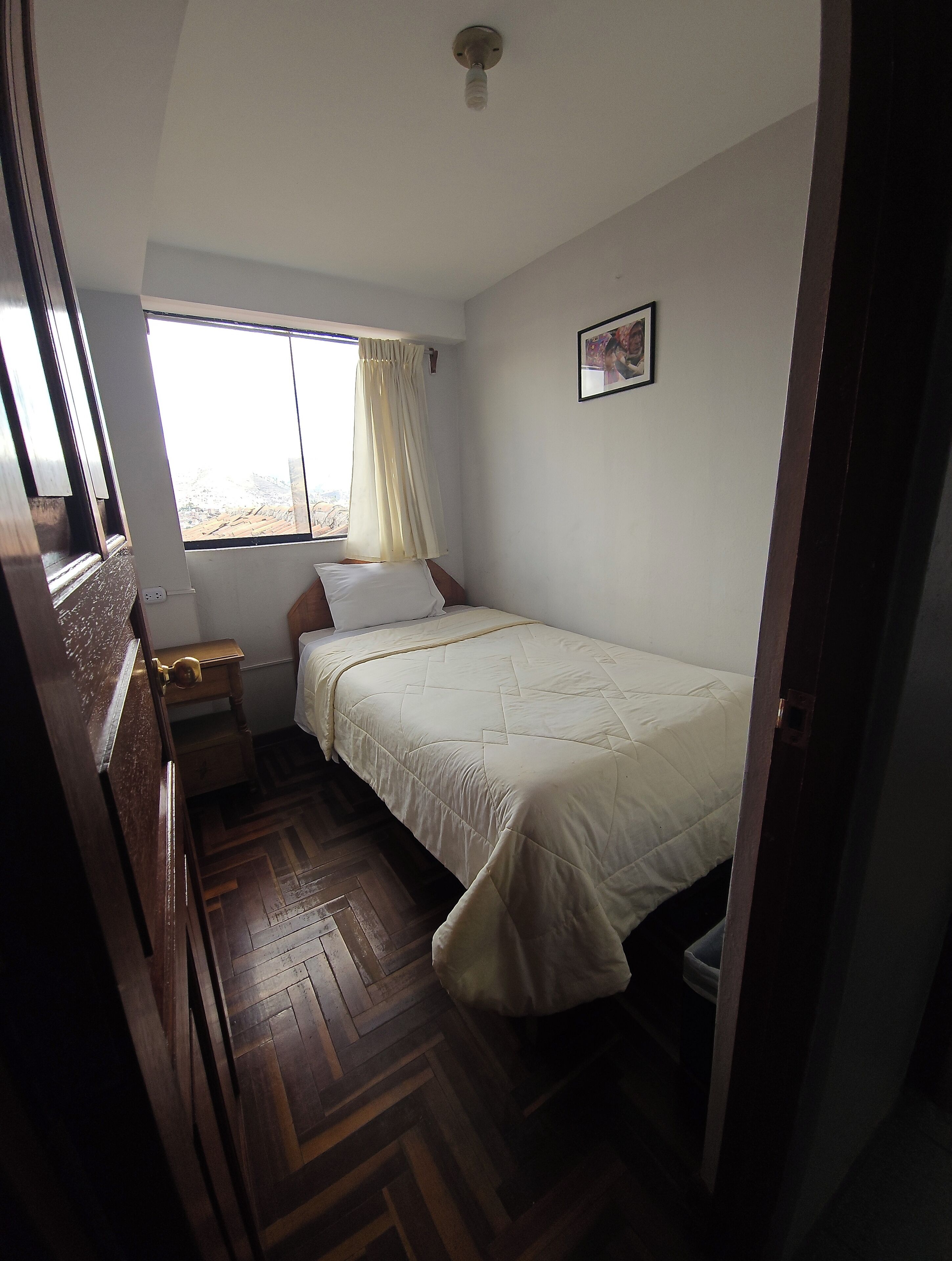 A bedroom with a bed, a window with curtains, a wooden dresser, and a small bedside table.