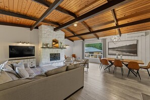 A modern living room with a stone fireplace, a large TV, and a dining area with wooden furniture. - Chalet Le Benjamin 4BDR - Pool - Jacuzzi (Mont-Tremblant)