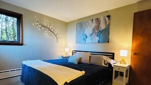 A bedroom with a large bed, two bedside tables, a wall-mounted abstract painting, a branch decoration, and a window with a view of trees.