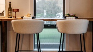 A room with a large window, two bar stools, a small table with a bottle and a candle, and a view of trees outside. - the tiny burrow ~ magical fleurieu escape (Aldinga)