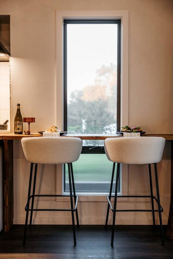 Una habitación con un ventanal grande, dos taburetes, una mesa pequeña con una botella y una vela, y vistas a árboles fuera.