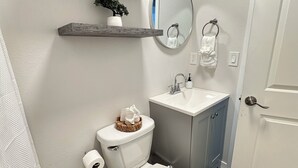 Un baño con inodoro, lavamanos y espejo. Hay toallas en un estante y una planta en maceta.