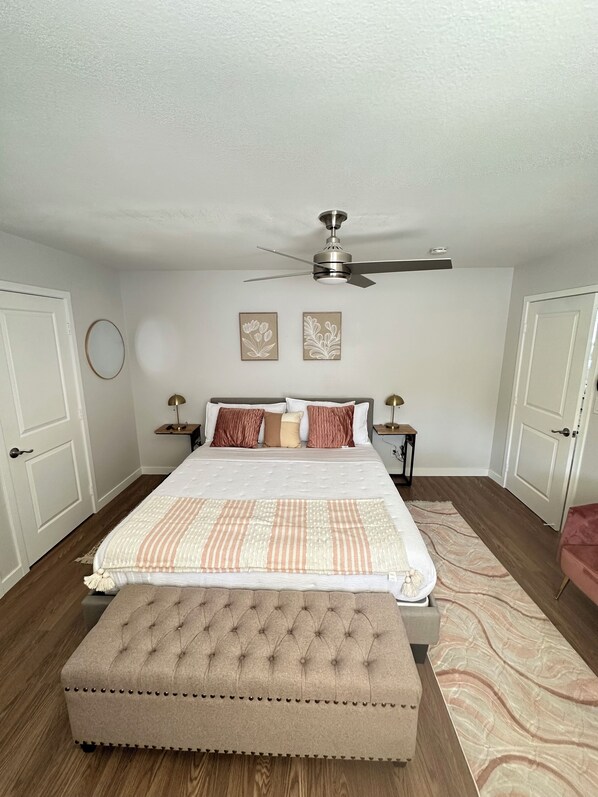 Un dormitorio con cama, ventilador de techo, dos obras de arte en la pared y piso de madera.