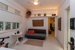 A living room with a sofa, a small table, a ceiling fan, and a wall-mounted shelf with decorative items. - Copacabana com Varanda – 3 min da praia (Rio de Janeiro)