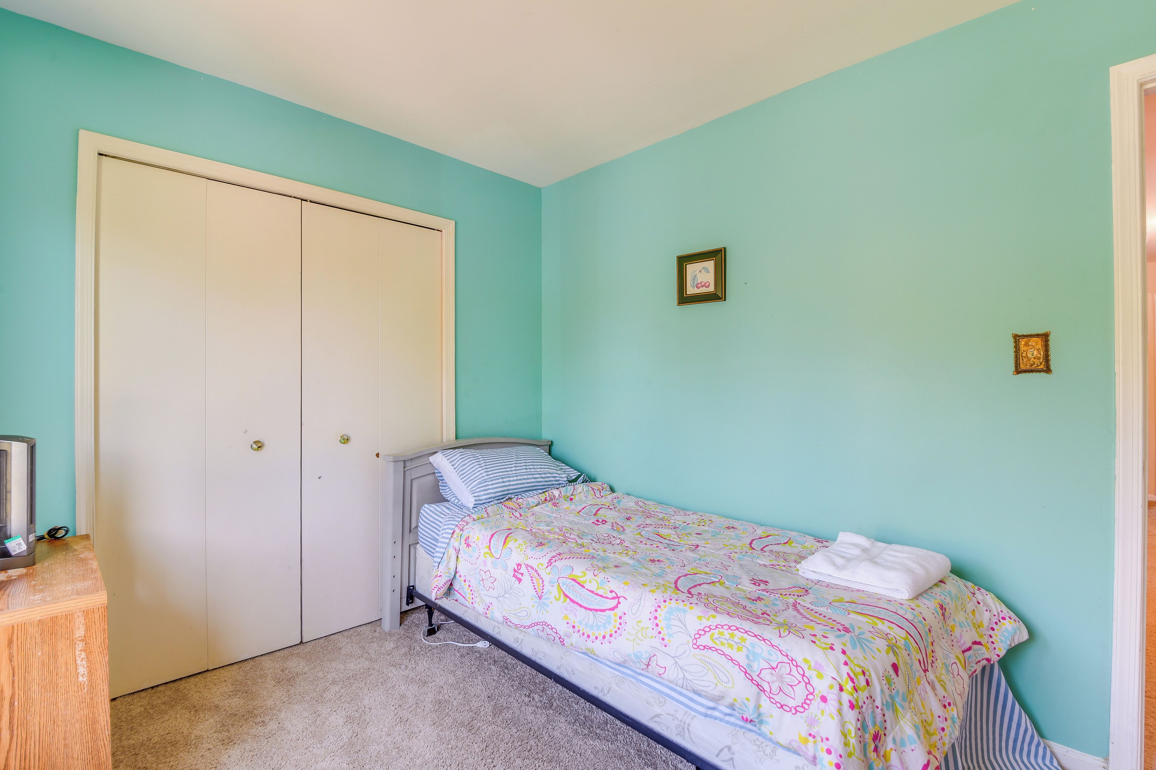 Ein Schlafzimmer mit türkisfarbenen Wänden, einem Bett mit farbenfroher Musterdecke, einem weißen Kleiderschrank und einem kleinen Schreibtisch mit einem Computer.