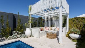 Een modern terras buiten met een witte pergola, een zwembad en een zithoek met kussens.