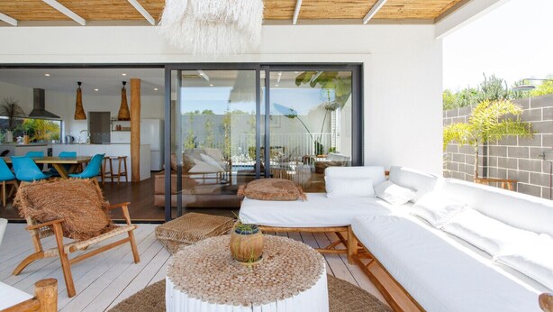 Een moderne woonkamer met een grote bank, een salontafel en rieten stoelen. Er is een glazen deur die leidt naar een balkon met uitzicht op de tuin.