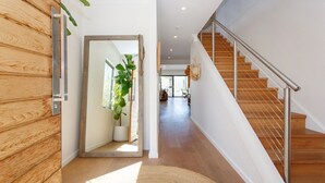 Een modern interieur met een houten vloer, een trap met metalen leuning en een grote spiegel die een kamer met een plant en een raam weerspiegelt.