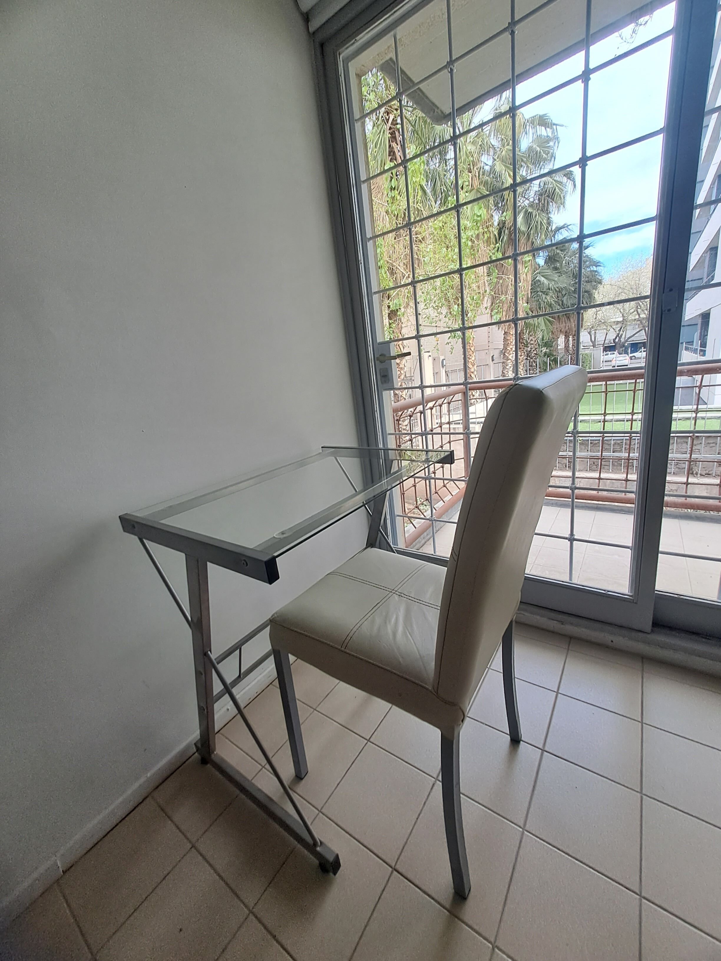 Una habitación con una mesa de vidrio y una silla, un ventanal con barrotes de metal y vistas a zonas verdes y edificios.