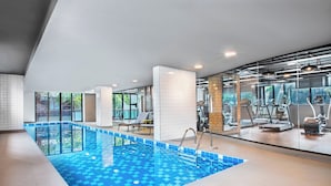 Una piscina interior con suelo azul a cuadros, rodeada de equipo de ejercicio y un muro de cristal con vista a zonas verdes.