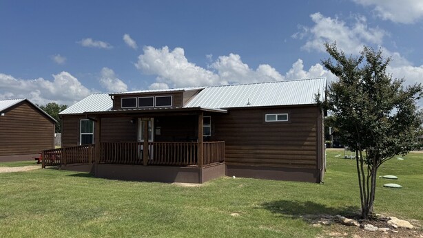 A two-story wooden cabin with a porch and a small tree in front. - Dinosaur Valley RV Park Cabin 6 (Glen Rose)