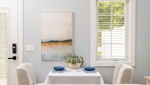 A room with a bed, a table set with blue bowls and wine glasses, a painting on the wall, and a window with blinds. - Cozy studio in Douglas with swimming pool and fireplace. (Douglas)