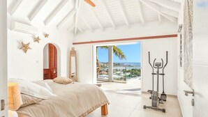 A bedroom with a bed, a workout area with exercise equipment, and a large window offering a view of the ocean.