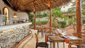 Un área de comedor al aire libre con paredes de piedra, mesas y sillas de madera, rodeada de follaje y un techo de paja.