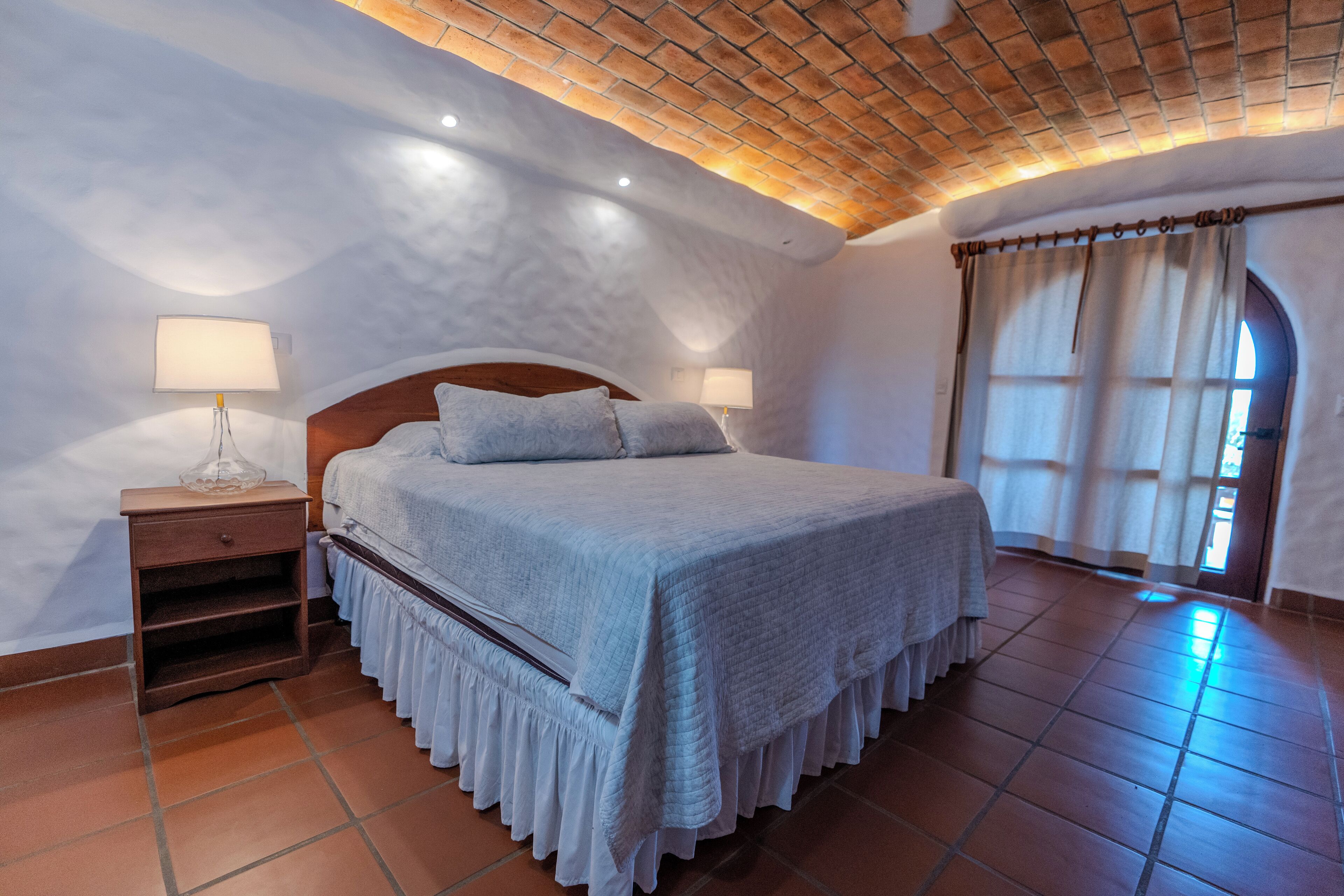 A bedroom with a bed, nightstands, a lamp, a window with curtains, and a tiled floor.