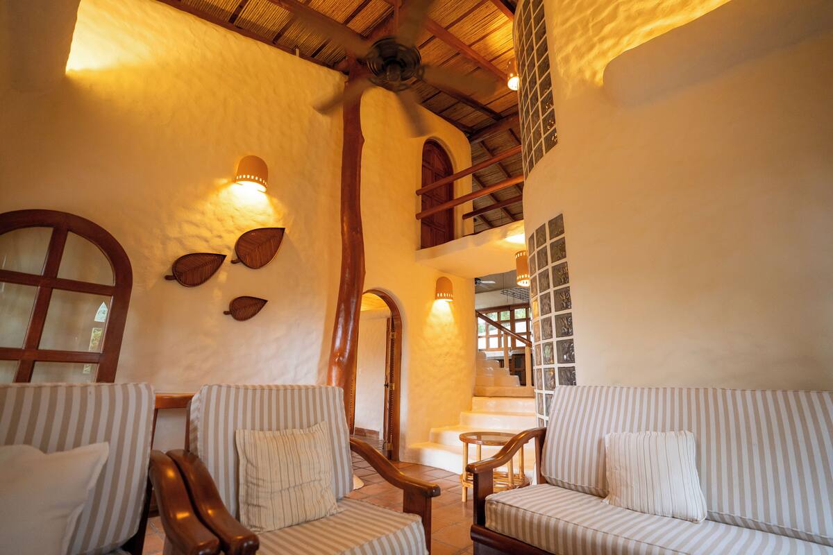 A room with striped sofas, a wooden ceiling fan, and a staircase with a glass railing.