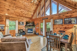 A log cabin interior with a fireplace, dining area, and large windows.